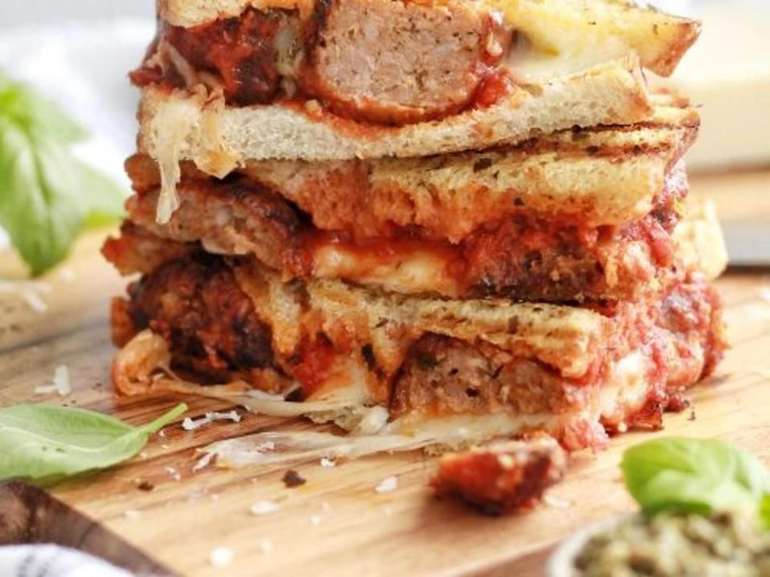 Close-up of a stack of meatball parmesan sandwiches with melted cheese, tomato sauce, and toasted bread, garnished with herbs on a wooden cutting board.