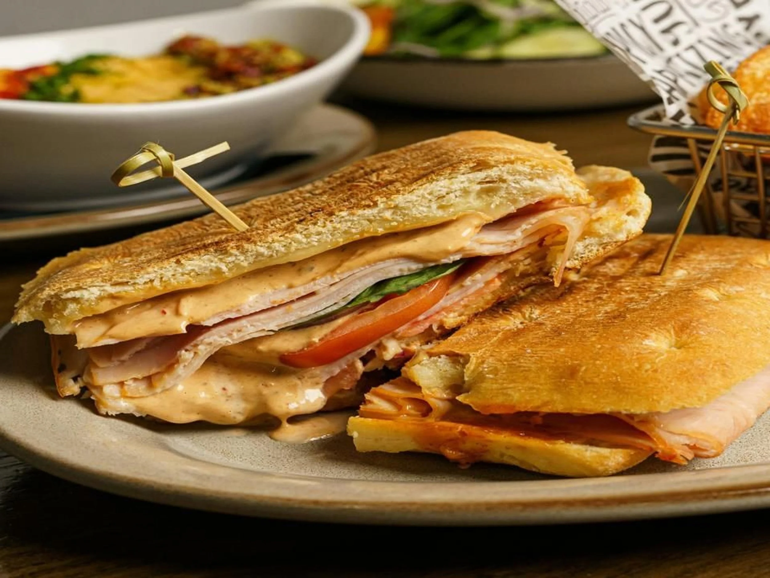 A sandwich on a plate with toasted bread, sliced turkey, tomato, lettuce, and cheese, with other dishes and plates in the background.