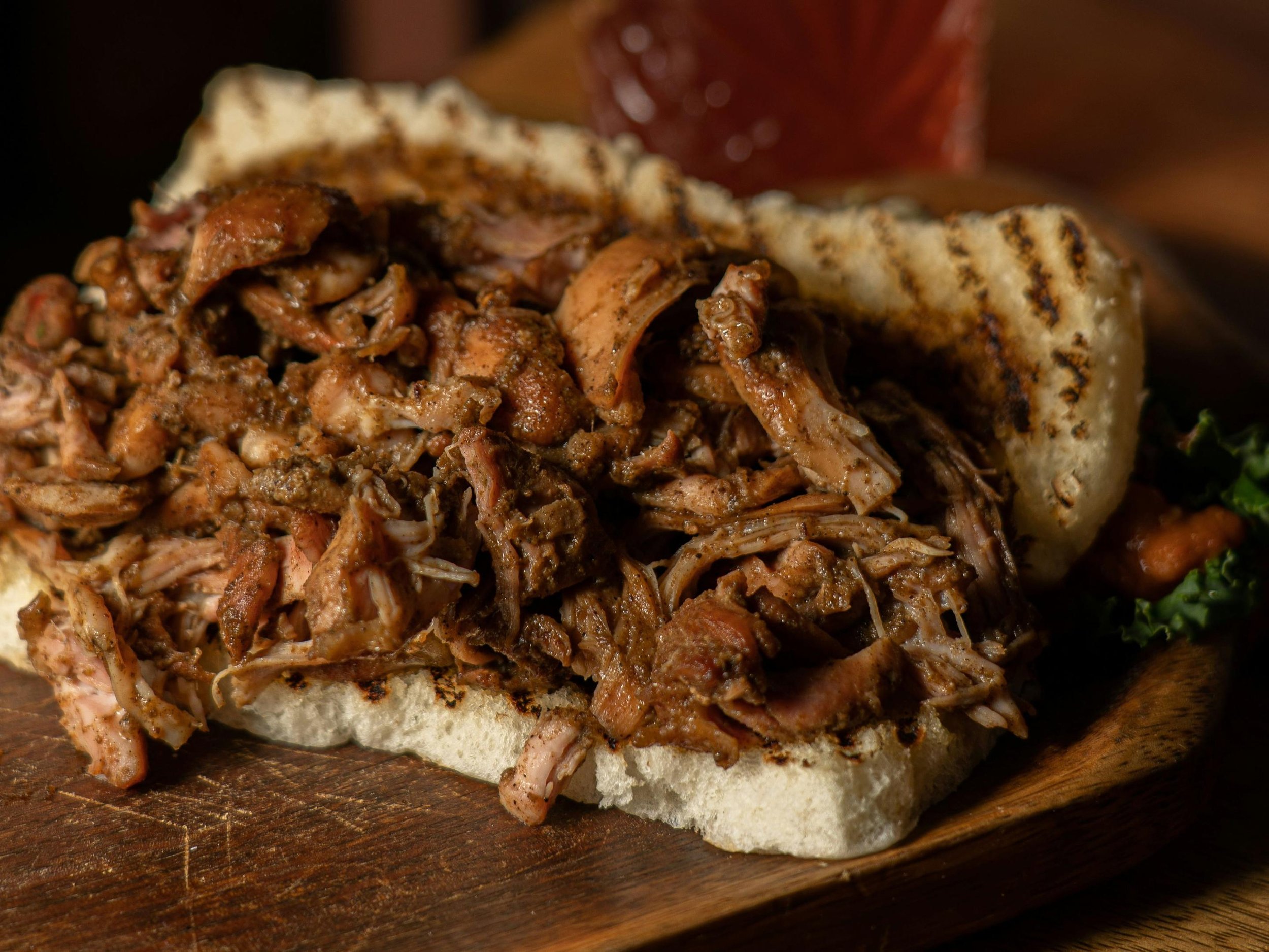 Pulled pork sandwich with shredded pork on a bun, served on a wooden board with a side of greens.