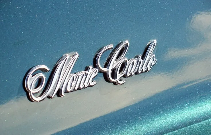 Close-up of a car's hood ornament with the words 'Monte Carlo' in chrome lettering on a blue painted surface.
