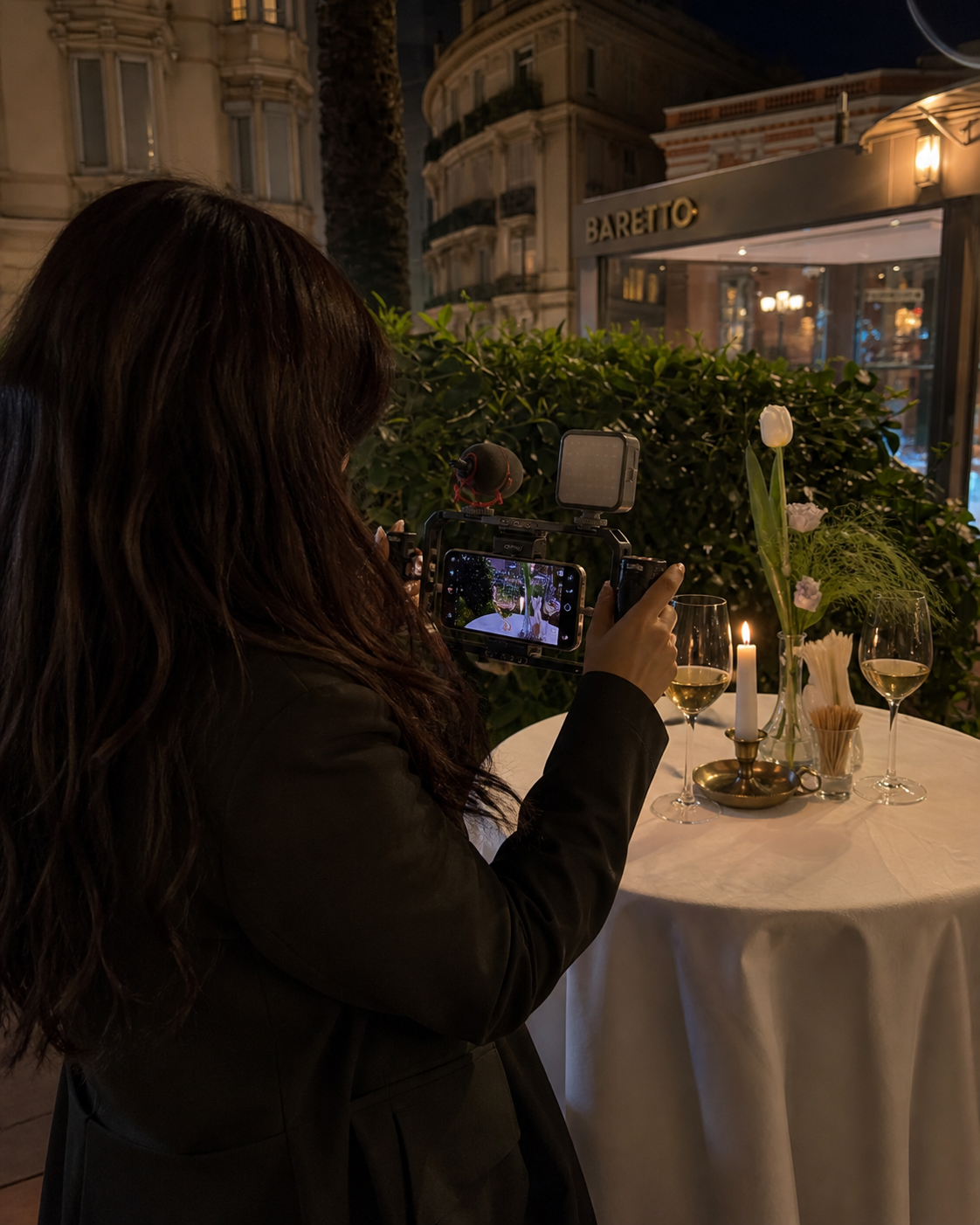 A woman with long dark hair is taking a photo with her smartphone at an outdoor dining table at night. The table has a white tablecloth, a lit candle, flowers, and glasses of water. A restaurant or cafe sign reads 'BARETO' in the background, with buildings and trees visible.