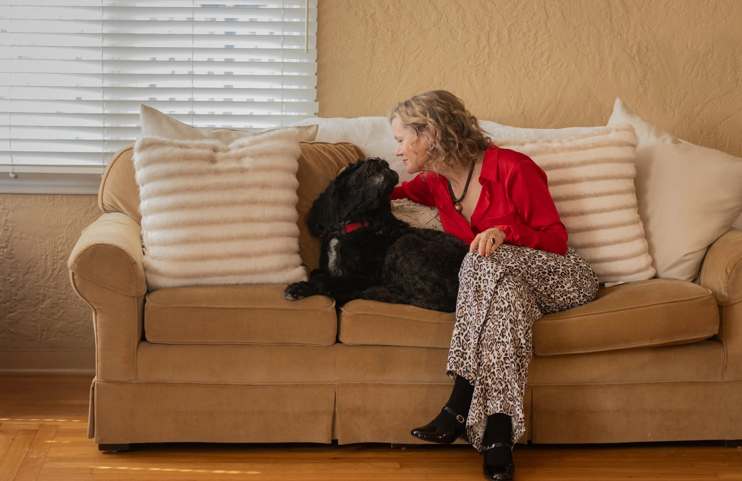 A woman with curly blonde hair wearing a red blouse and leopard print pants sitting on a beige sofa with multiple pillows, looking at a black dog with curly fur, near a window with white blinds.