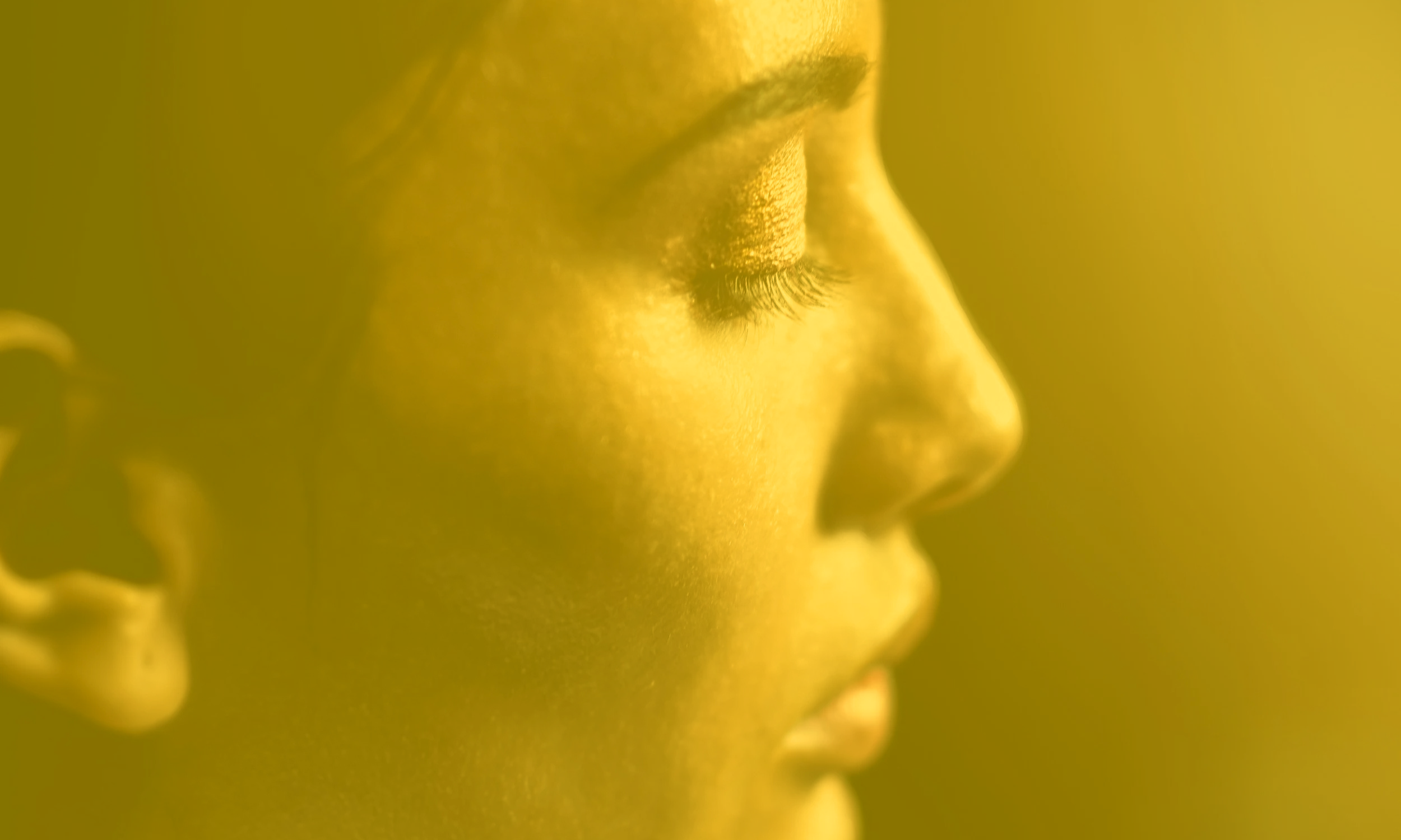 Close-up side profile of a person's face, likely a woman, with closed eyes and long eyelashes, bathed in warm yellow lighting.