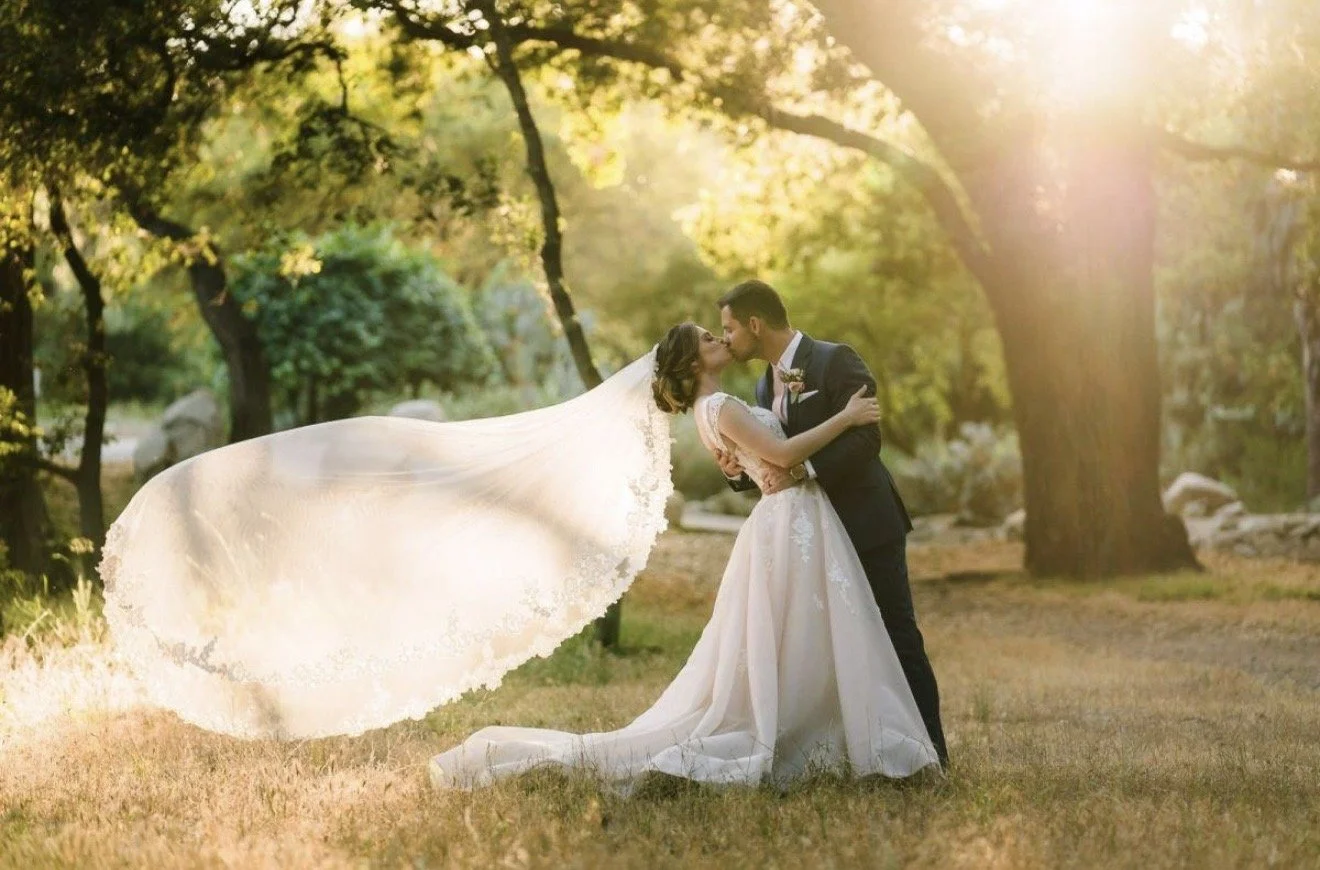 A bride and groom kiss in a park during sunset, surrounded by trees with golden leaves.