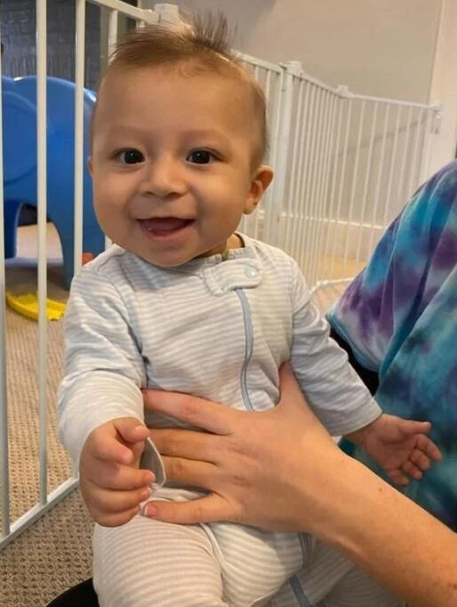 A smiling baby sitting on an adult's lap, with a baby gate and blue chair in the background.