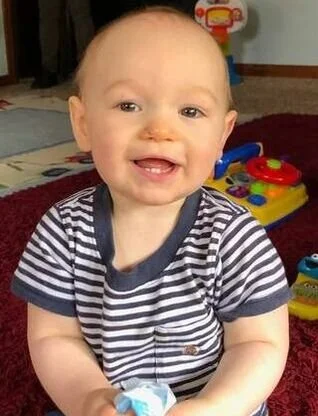 Smiling baby sitting on a red carpet with toys in the background.