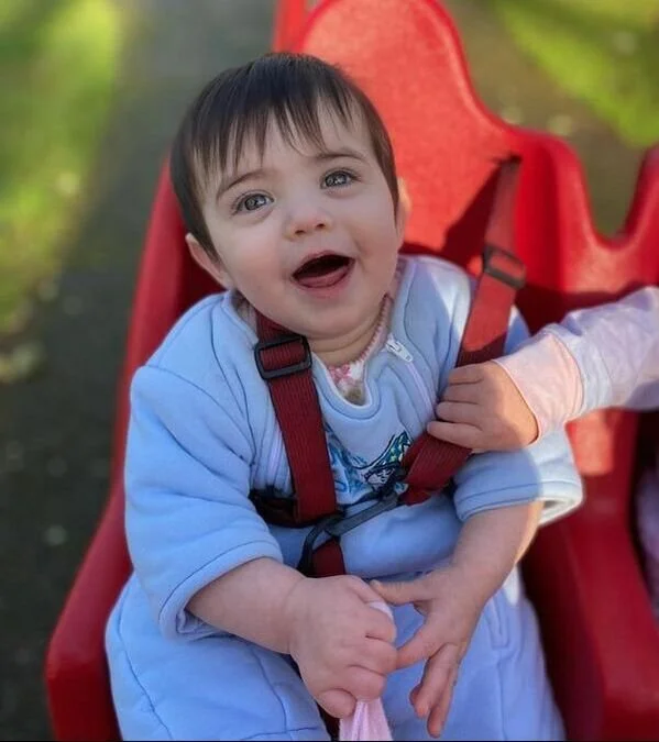 A smiling toddler with dark hair and blue eyes sitting in a red outdoor swing, wearing a light blue outfit, with a hand helping to secure the harness.
