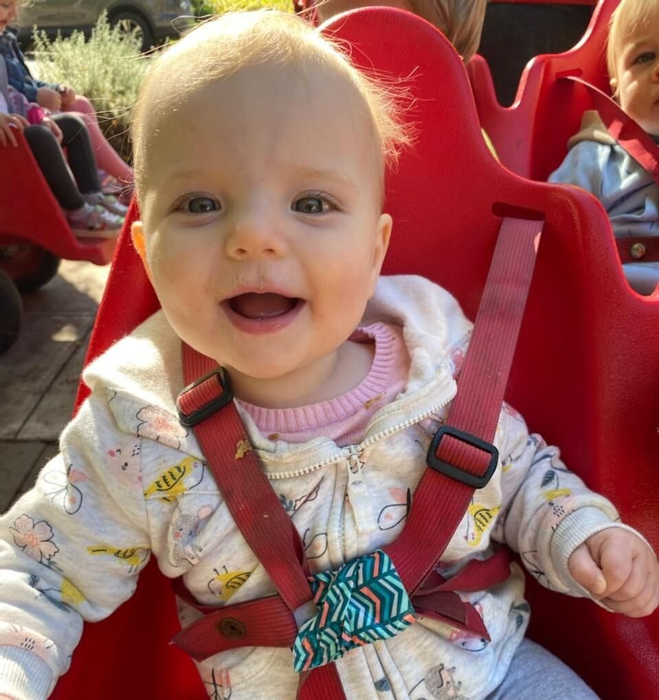 A smiling baby sitting in a red stroller outdoors, wearing a light-colored jacket with a colorful pattern and a striped bow tie, with other children visible in the background.