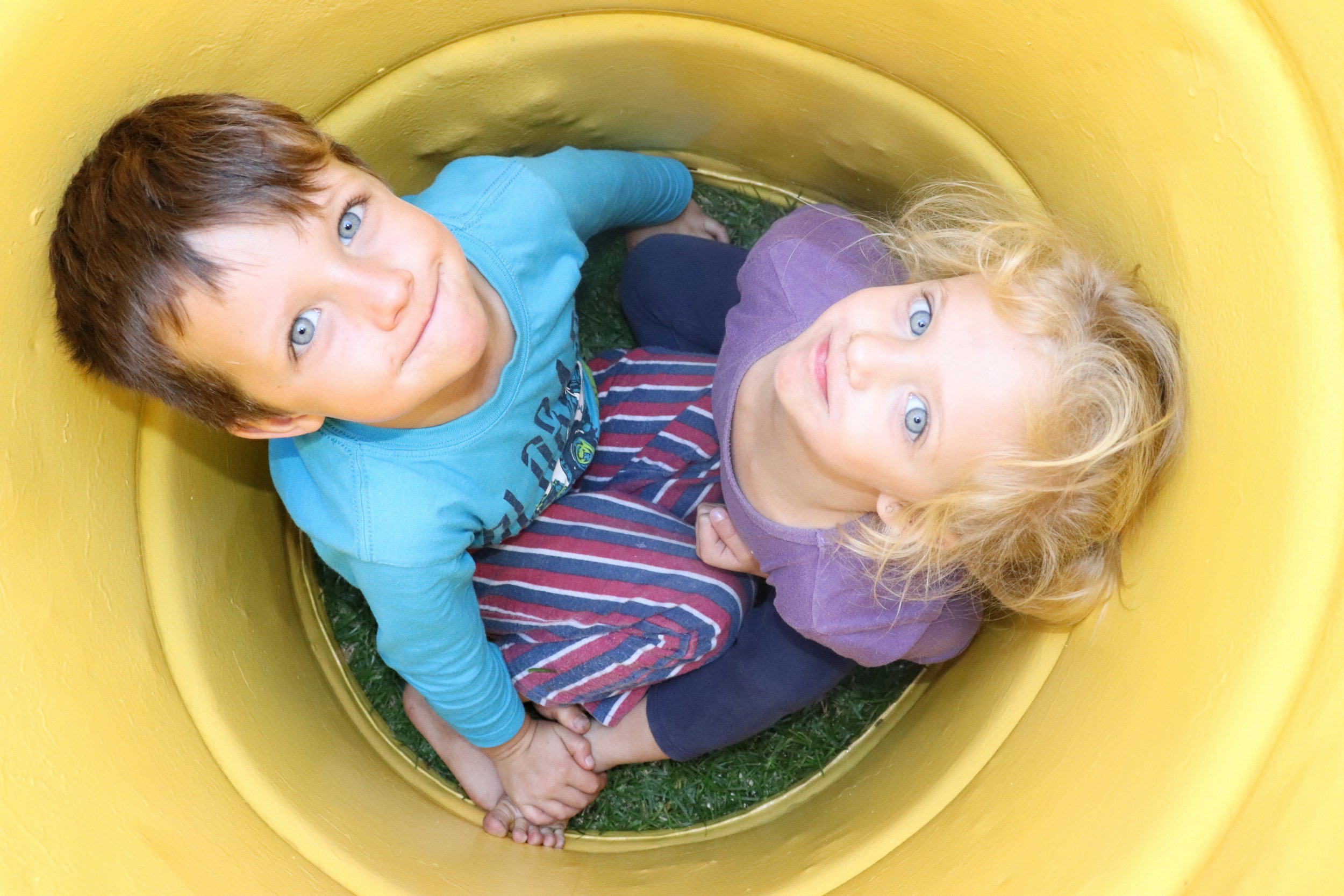 Two children, one boy with light brown hair and blue eyes wearing a light blue shirt, and one girl with curly blonde hair and blue eyes wearing a purple shirt, are inside a yellow tunnel slide, sitting on the grass. They are facing the camera and smiling gently.