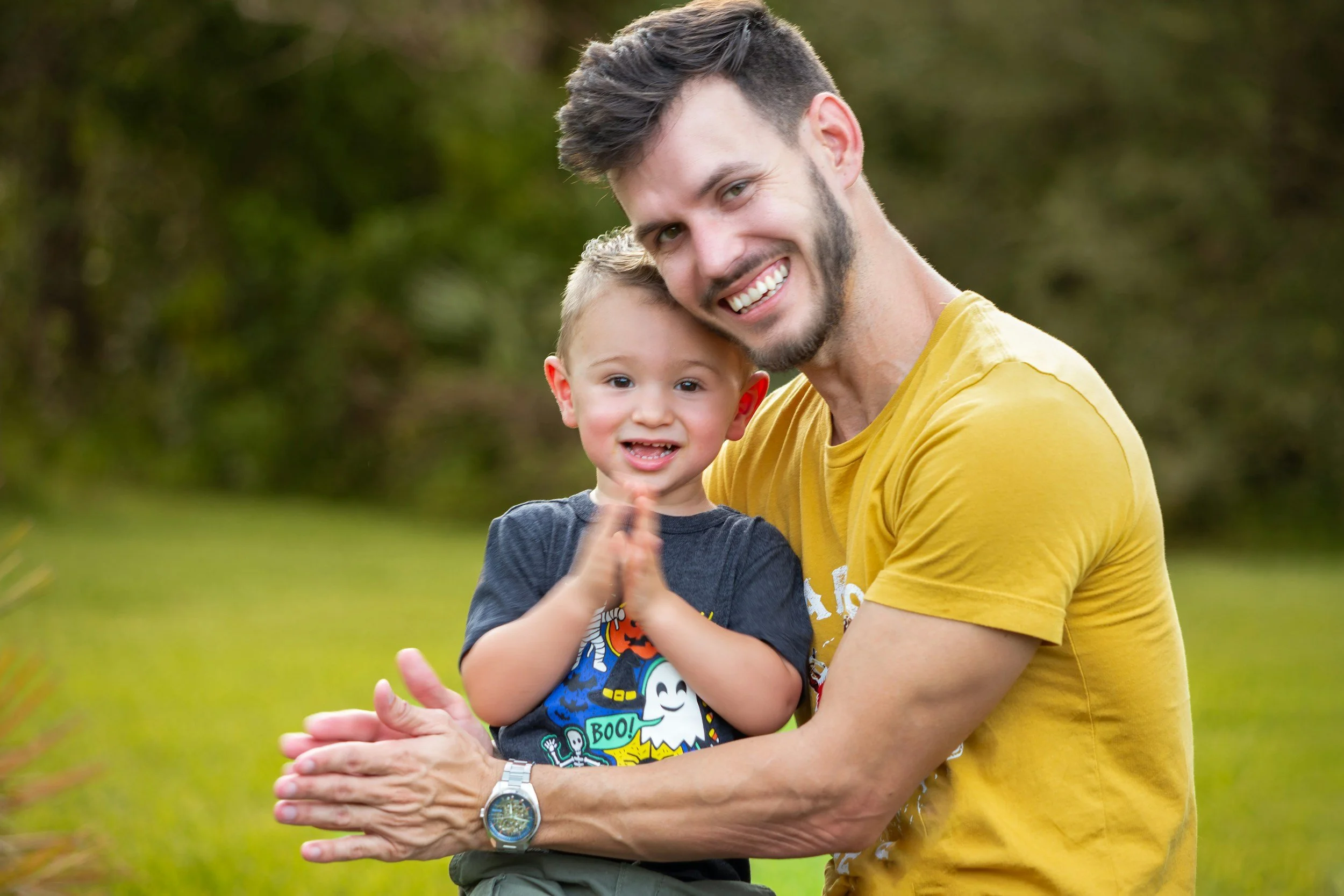 A man with dark hair, a beard, and a yellow T-shirt smiling and holding a young boy with short blonde hair and gray T-shirt with Halloween-themed design outdoors with blurred green trees in the background.