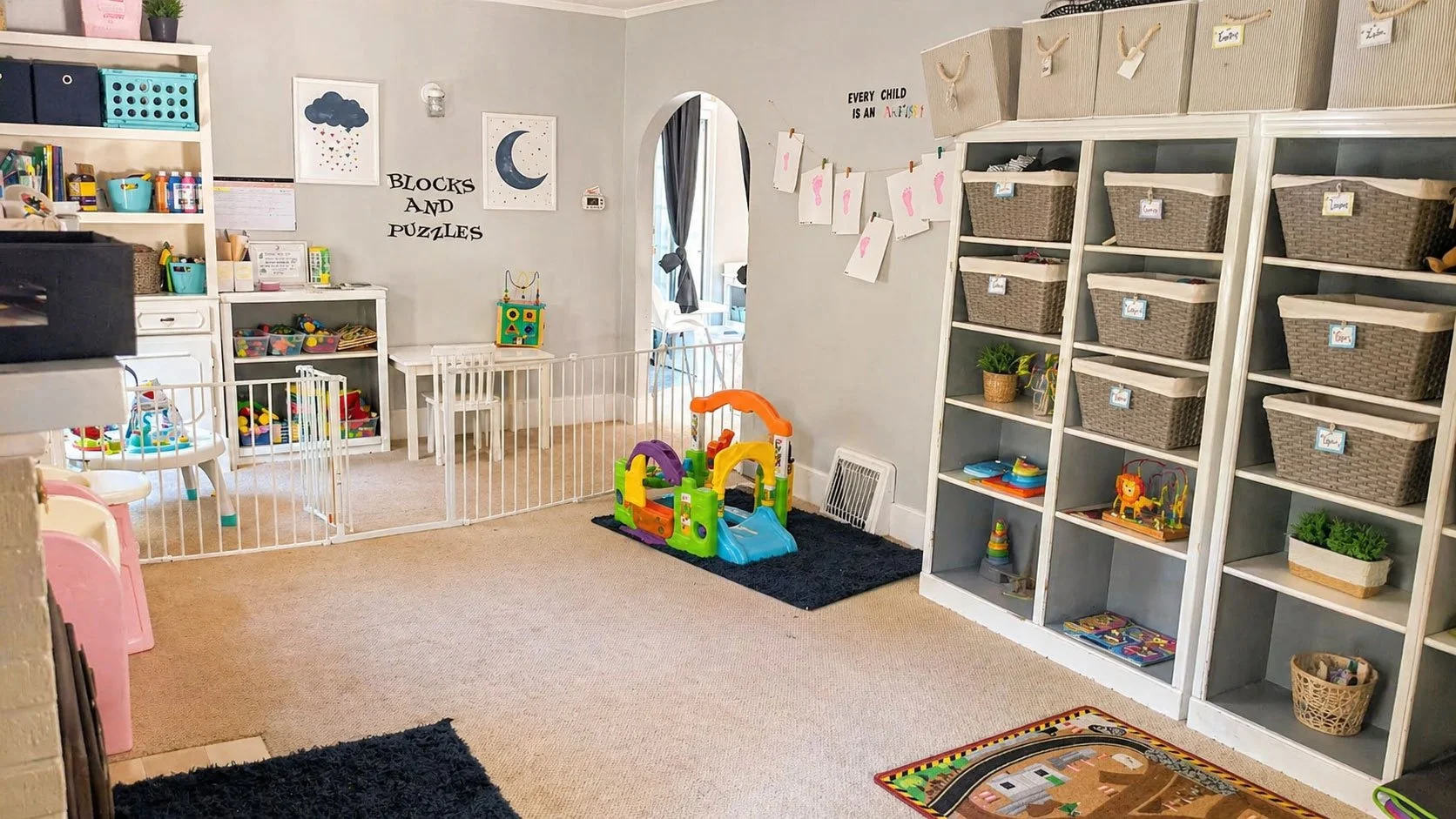 A colorful children's playroom with toys, books, and storage bins, decorated with moon and cloud wall art, and with a child-safe gate. There are shelves filled with toys, a small playhouse, and artwork on the walls.