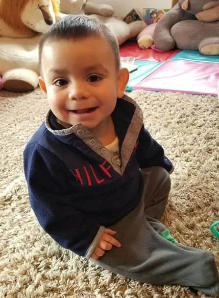Young boy sitting on a beige carpet in a playroom, smiling at the camera, with plush toys in the background.