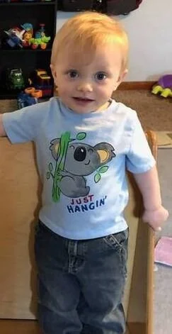 A young boy with blonde hair and blue eyes standing indoors, wearing a light blue T-shirt with a koala cartoon and the words "Just Hangin'" on it. There's furniture and toys in the background.