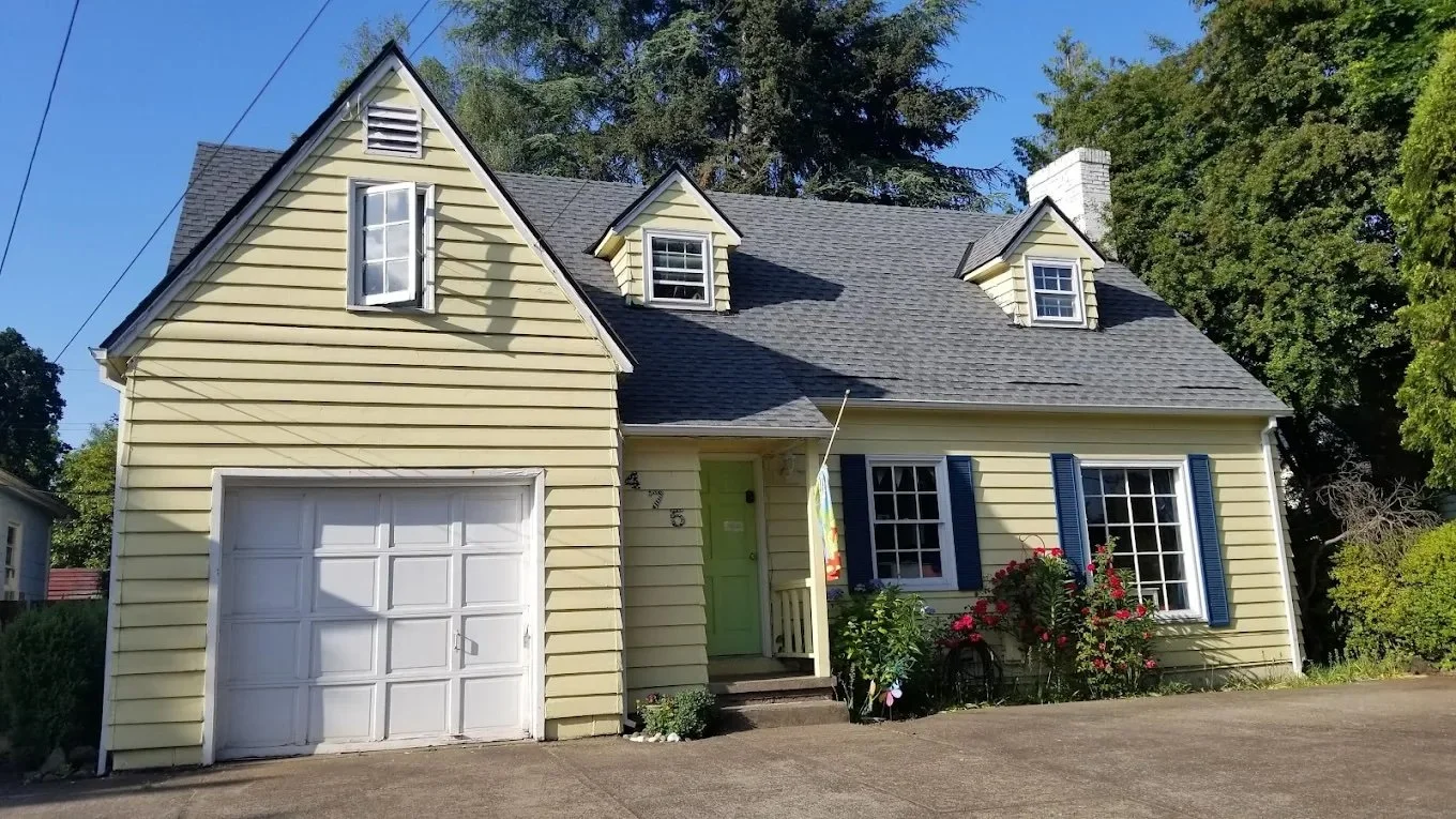 A yellow house with blue shutters, a garage, and a light green front door. There are flowers and bushes in the front yard, and the house has a gray shingled roof with three dormer windows. Trees surround the house, and it is daytime with a clear blue sky.