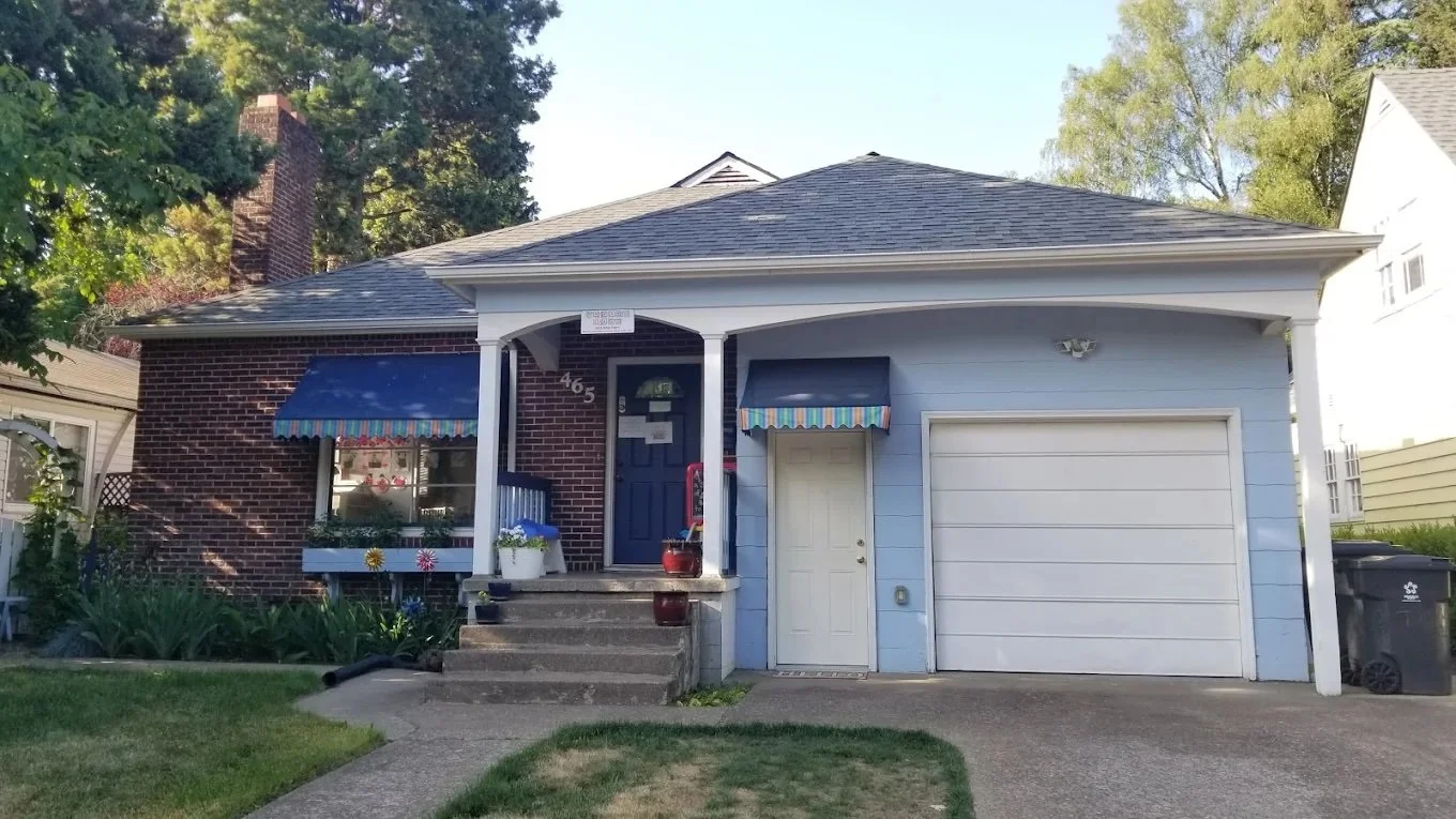 A house with a brick and light blue exterior, front stairs, a garage door, and a small front porch with striped awnings and plants.