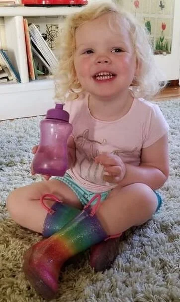 A young girl with curly blonde hair sitting on a gray carpet, holding a purple sippy cup, wearing rainbow-colored rain boots and a pink shirt, smiling at the camera.