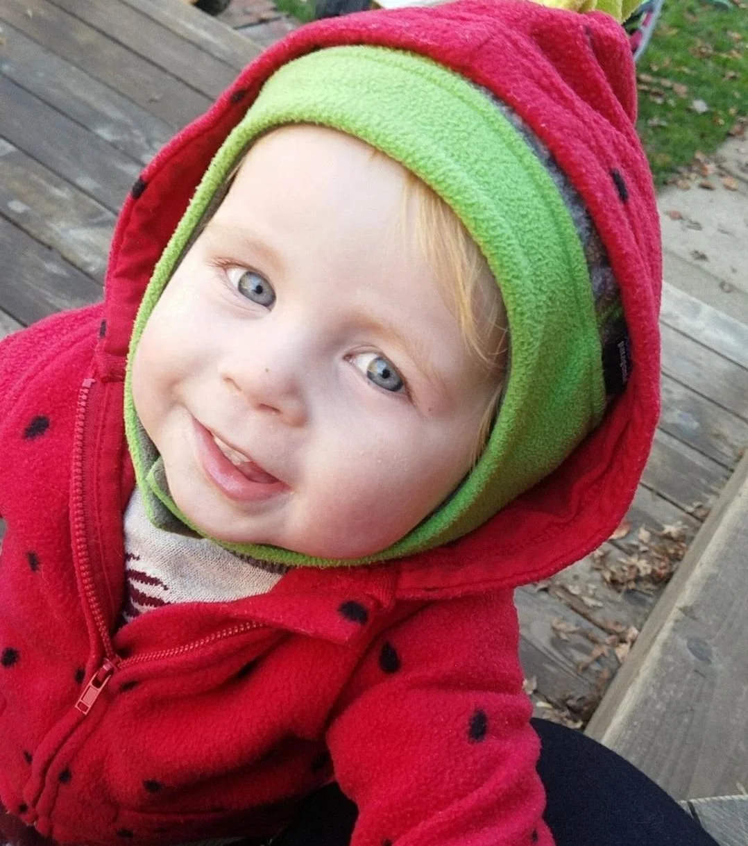 Close-up of a smiling toddler with blue eyes, wearing a red fleece jacket with black dots and a green and red hood, outdoors on a wooden platform.