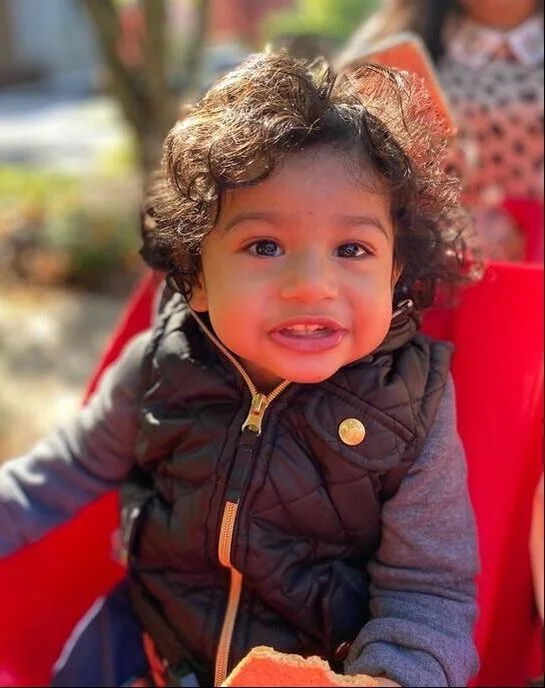 A young child with curly hair and a bright expression sitting outdoors, wearing a black quilted vest over a gray long-sleeve shirt, surrounded by greenery.