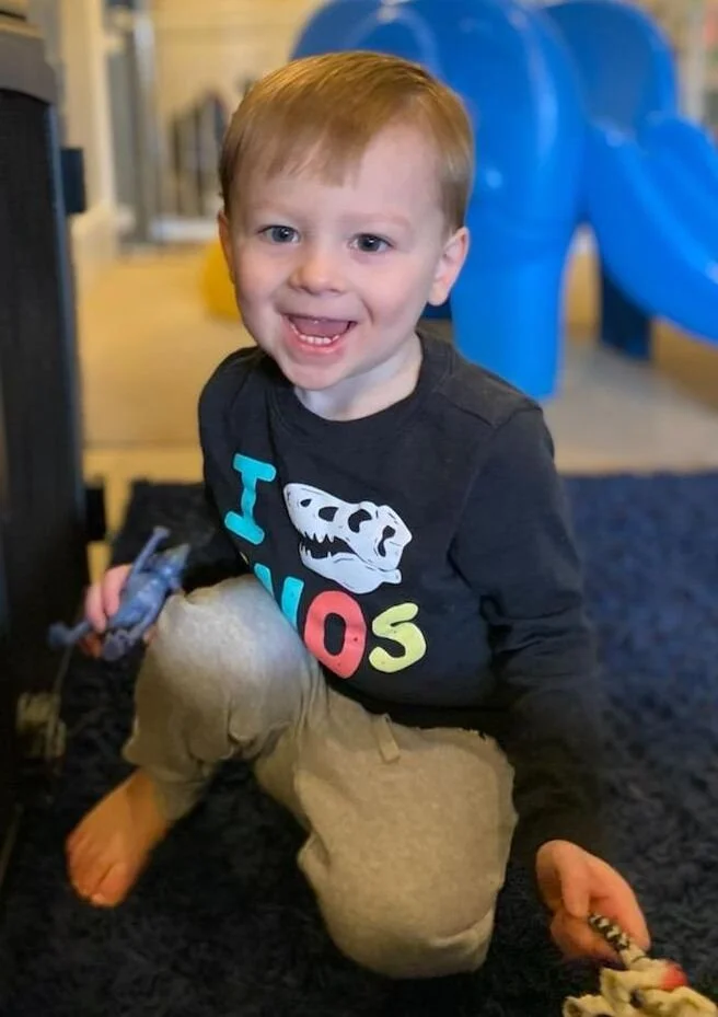 A young boy with red hair, wearing a black long-sleeve shirt with colorful text and a dinosaur graphic, is sitting on a black carpeted floor indoors, holding toys in both hands, smiling at the camera, with a blue plastic play structure in the backgro