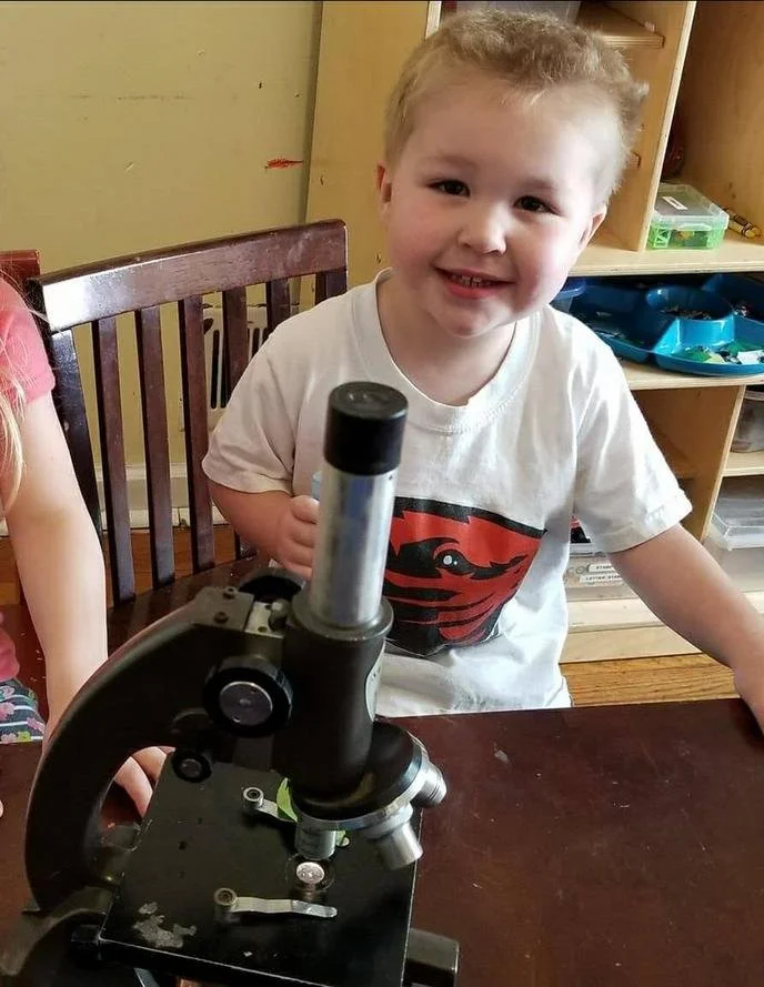 A young boy smiling and holding a microscope in a classroom or play area.
