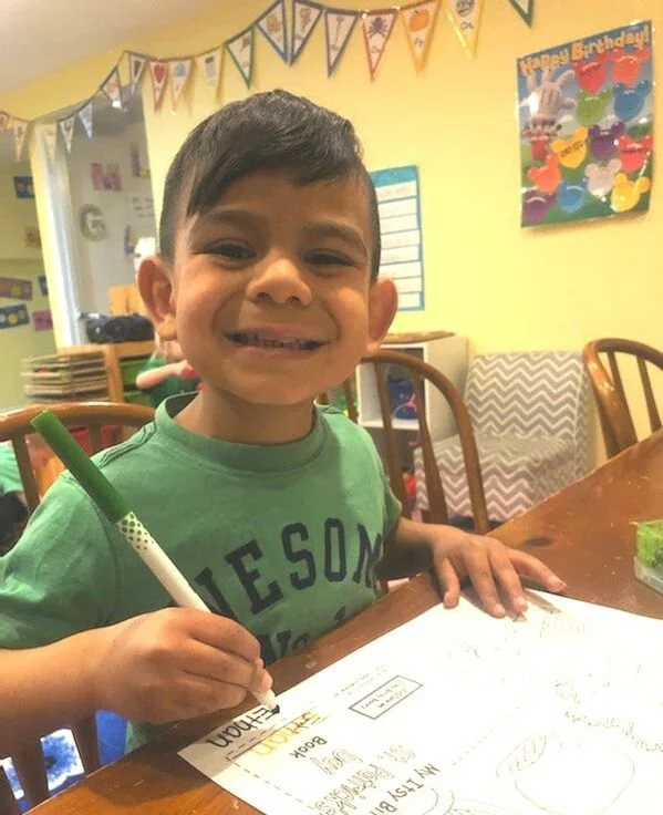 A young boy with dark hair smiling at the camera, sitting at a table in a classroom decorated with colorful banners and birthday poster.