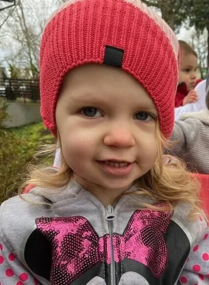 Close-up of a young girl wearing a red knit beanie and a gray jacket with pink and black details, outdoors with trees and another child in the background.
