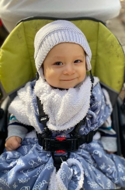 A smiling young child sitting in a green and black stroller, wearing a white knit hat, a white fluffy scarf, and a blue patterned snowsuit.