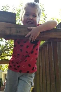 Smiling young girl with blonde hair leaning on a wooden railing outdoors, wearing a red shirt with black polka dots and beige pants.