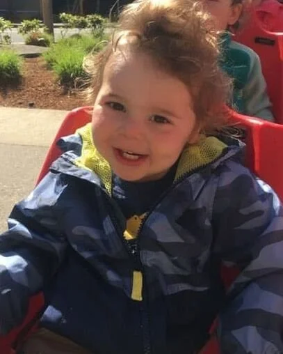 Smiling young child with curly hair sitting outdoors in a red stroller, wearing a blue camouflage jacket.