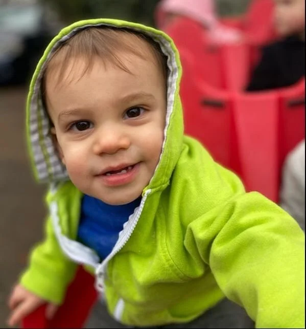 Close-up of a smiling young child wearing a bright green hoodie with striped lining, outdoors with a red stroller in the background.