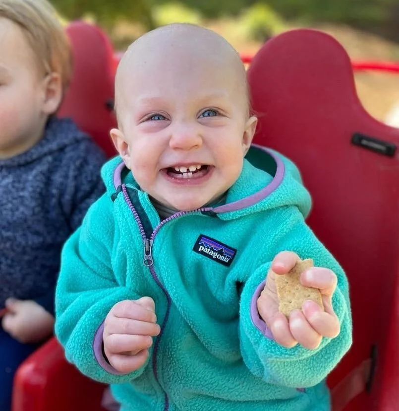 A smiling toddler with light skin and blue eyes sitting outdoors, holding a snack, wearing a turquoise Patagonia fleece jacket, with another child partially visible beside him, and a red seat in the background.