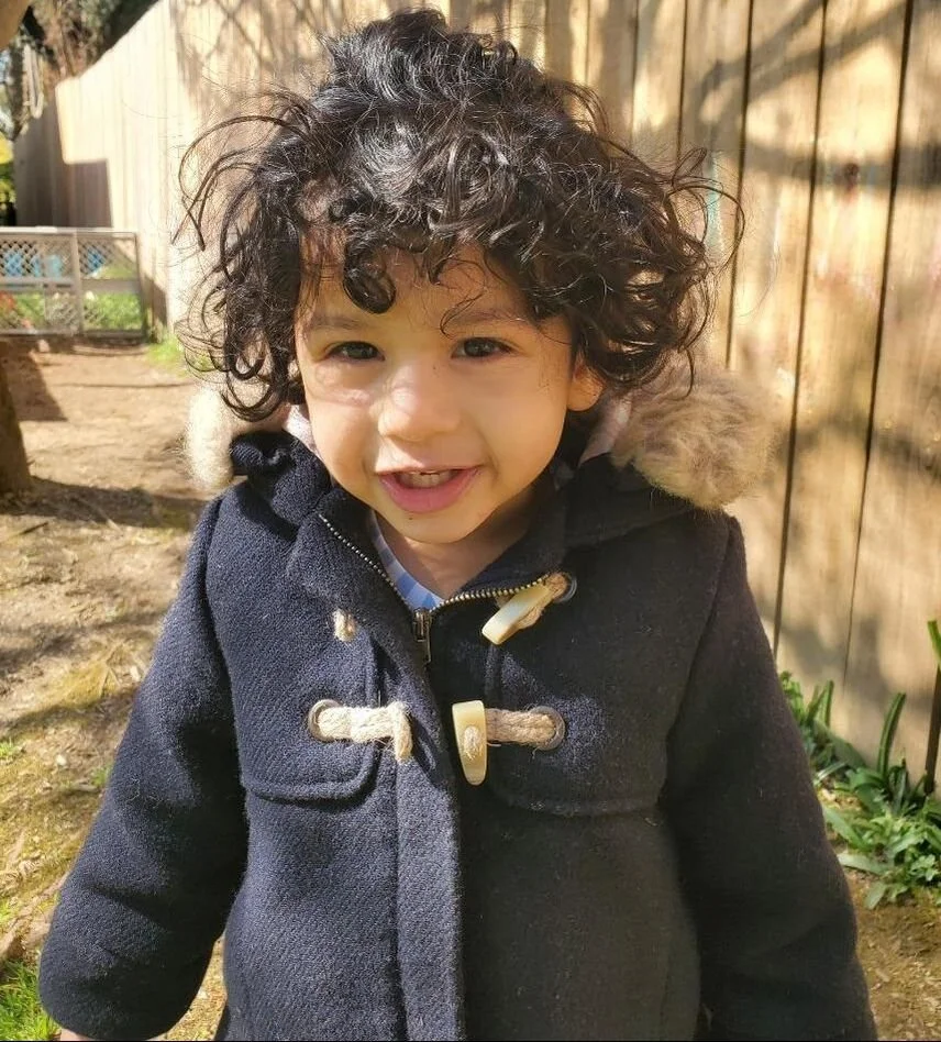 A young child with curly dark hair wearing a black coat with a fur-lined hood, smiling outdoors in sunlight with a wooden fence and plants in the background.