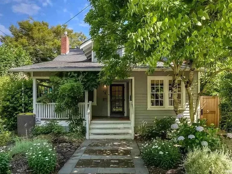A cozy house with a small front porch and a pathway leading to the front door, surrounded by lush green plants and trees, under a partly cloudy sky.