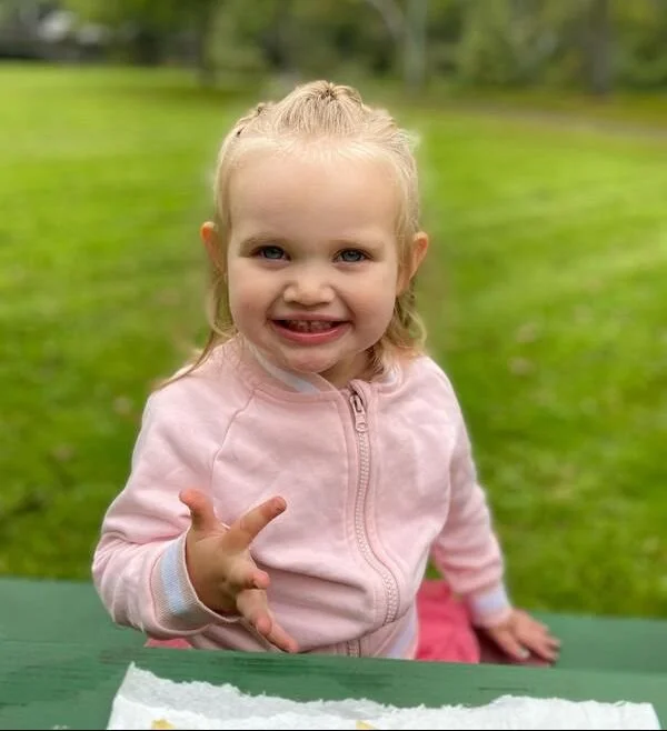 A young girl with blonde hair in a ponytail, smiling and making a peace sign with her right hand, sitting outdoors at a green table.