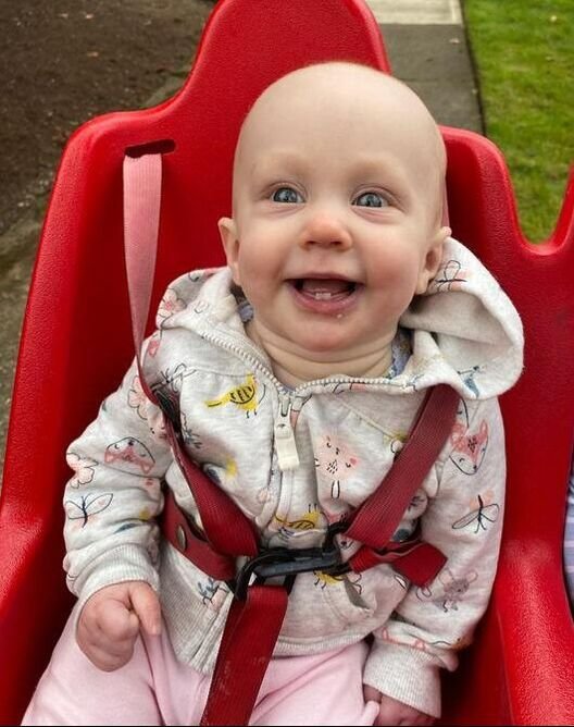 A happy baby girl with blue eyes and a big smile, sitting in a red swing or seat outdoors, wearing a light-colored hoodie with colorful prints and light pink pants.