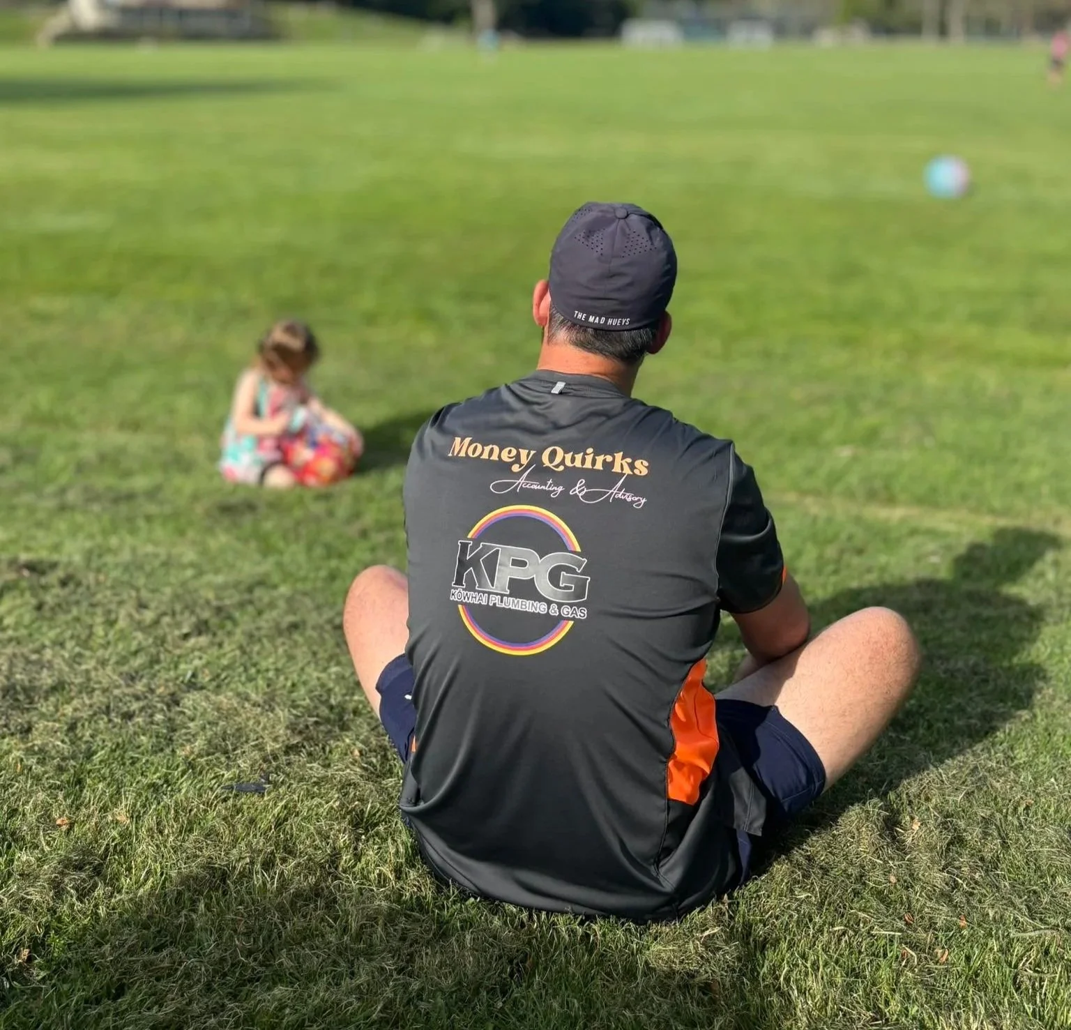 A man wearing a black and orange sports shirt with logos sits cross-legged on the grass, facing away from the camera, with a young girl sitting on the grass in front of him in the background.