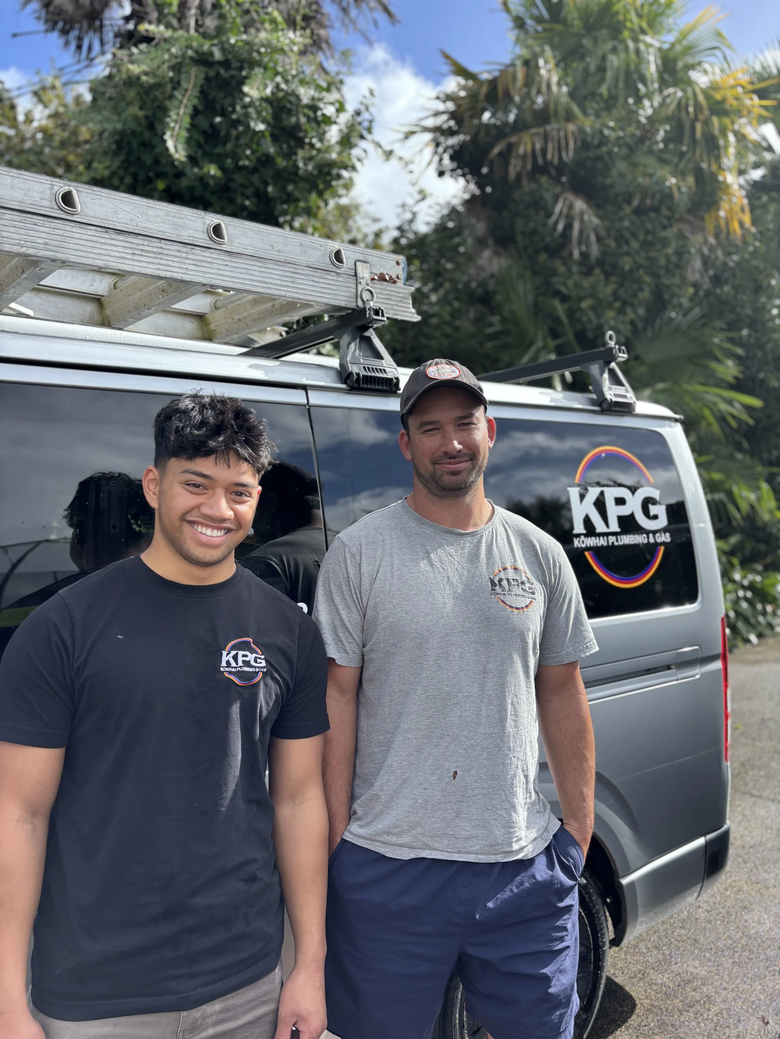 Two men standing in front of a van with a ladder on top, wearing T-shirts with a company logo, outdoors with trees and a blue sky in the background.