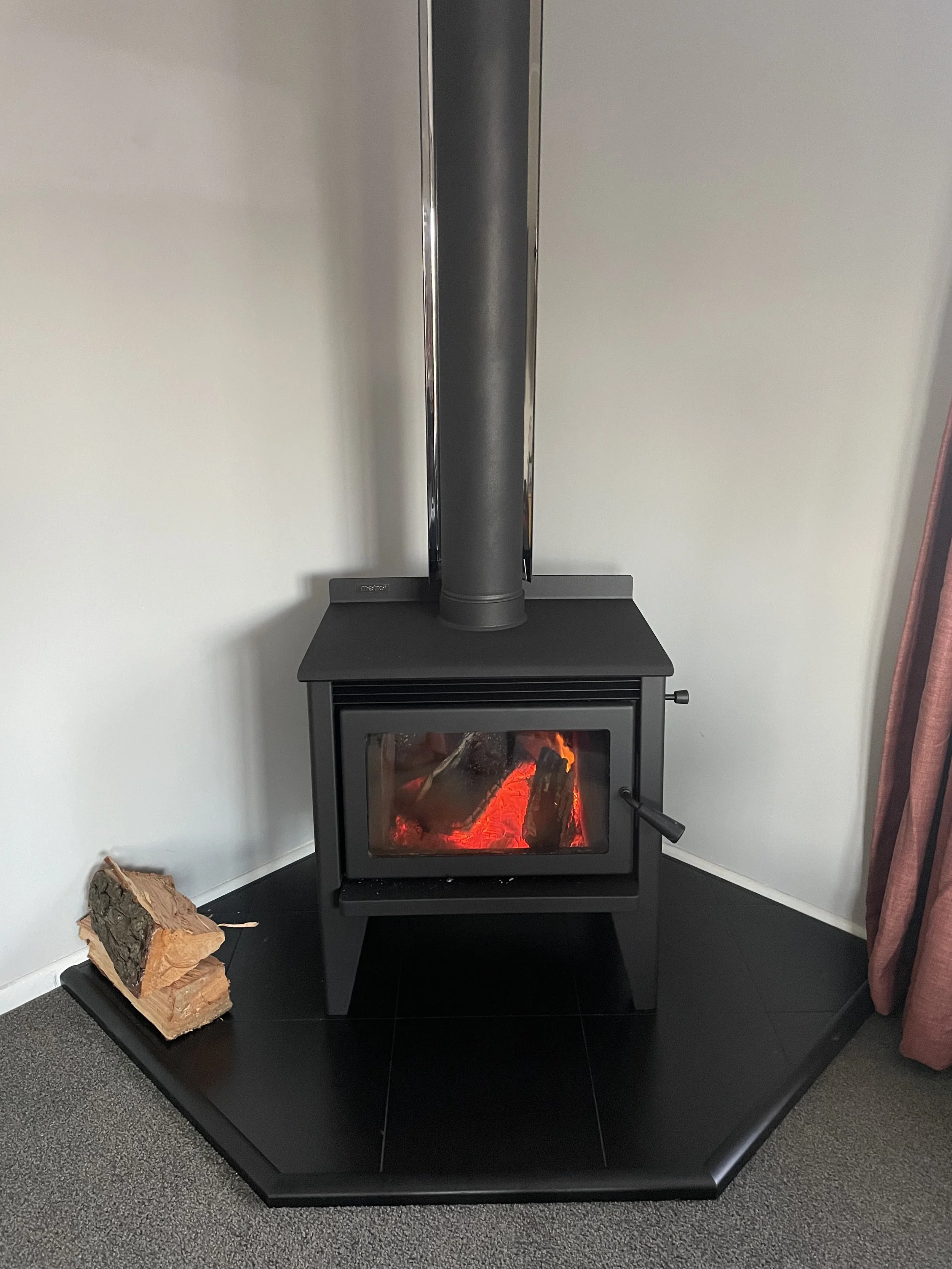 A black wood stove with a glass door showing glowing orange embers inside. There is a pile of chopped wood on a black table next to the stove, and a metal pipe extending from the stove to the ceiling. The room has white walls and gray carpet.