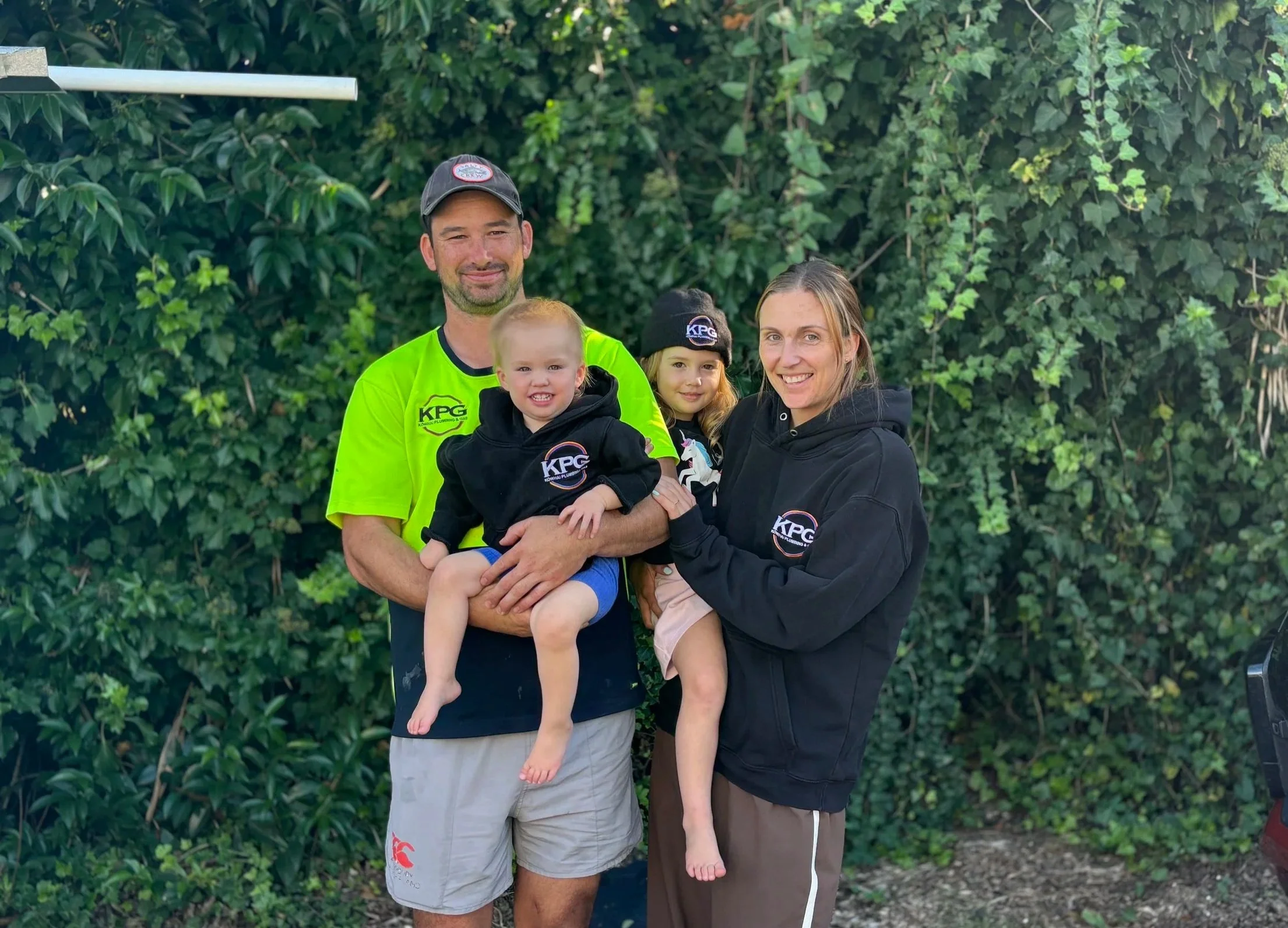 A family of four standing outdoors in front of green foliage, smiling. The man is holding a young girl, the woman is holding a baby, and a teenage girl is next to them, all dressed casually.