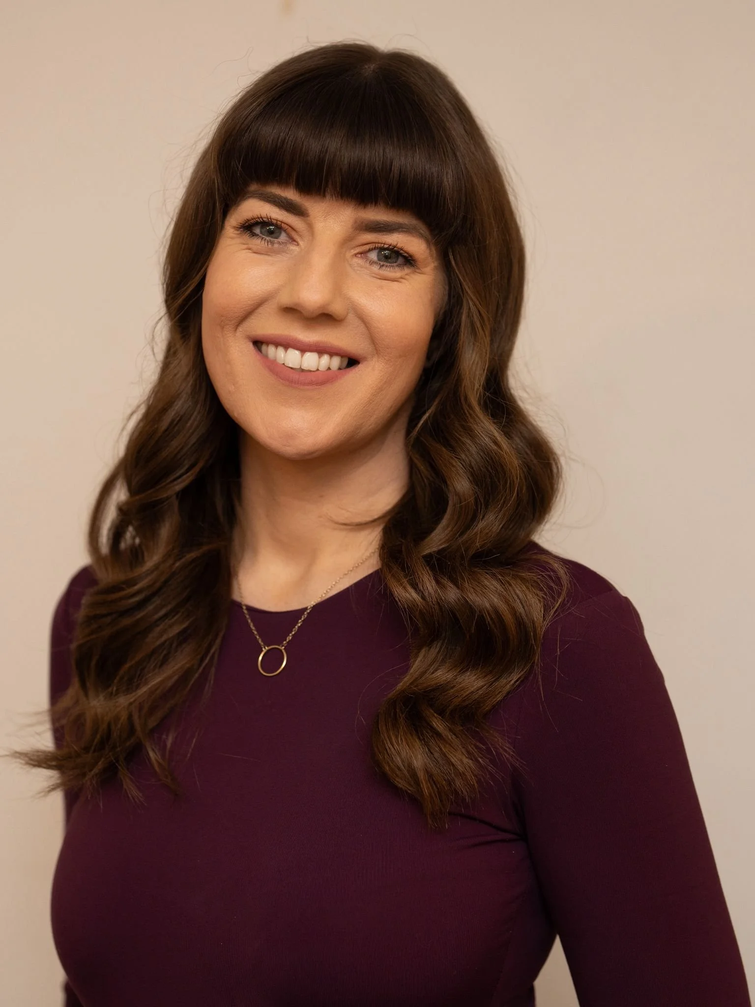Portrait of a woman with wavy brown hair and bangs, wearing a dark purple top and a gold necklace with a circular pendant, smiling against a neutral background.