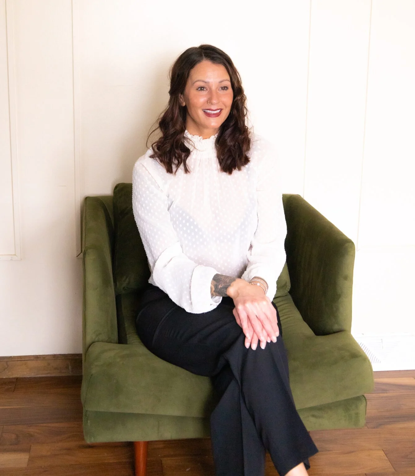 A woman with dark brown hair, smiling, sitting on a green couch against a plain white wall, wearing a white blouse with textured polka dots and black pants.