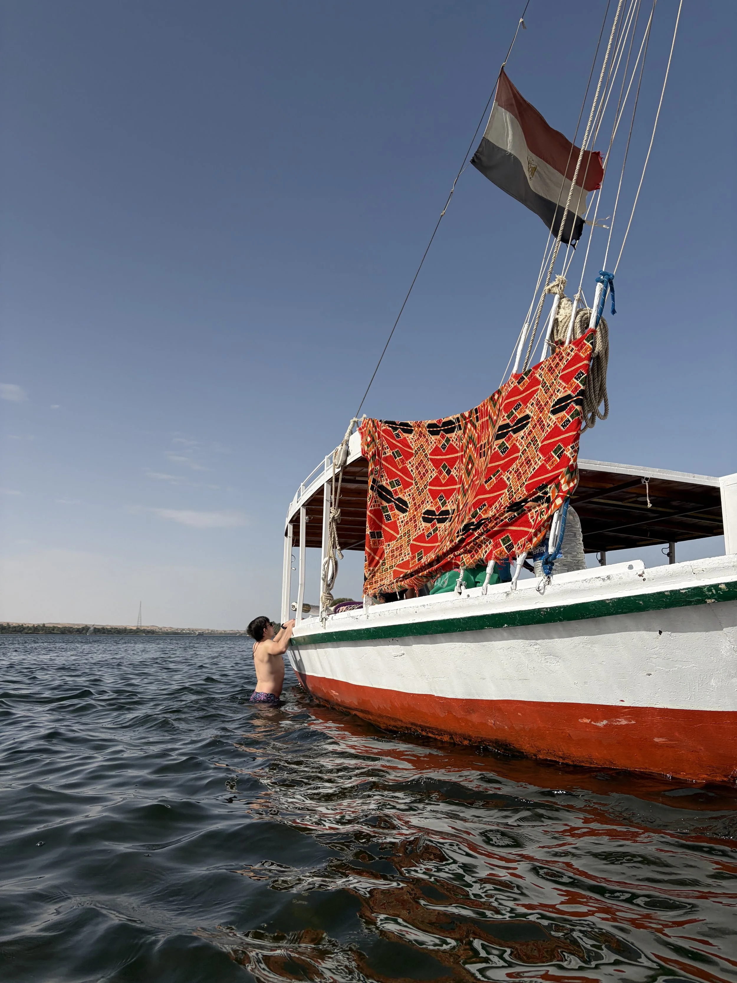 A child in swim trunks climbing onto a boat while in the water. The boat has a red, black, and white flag and a brightly colored blanket hanging from its side.