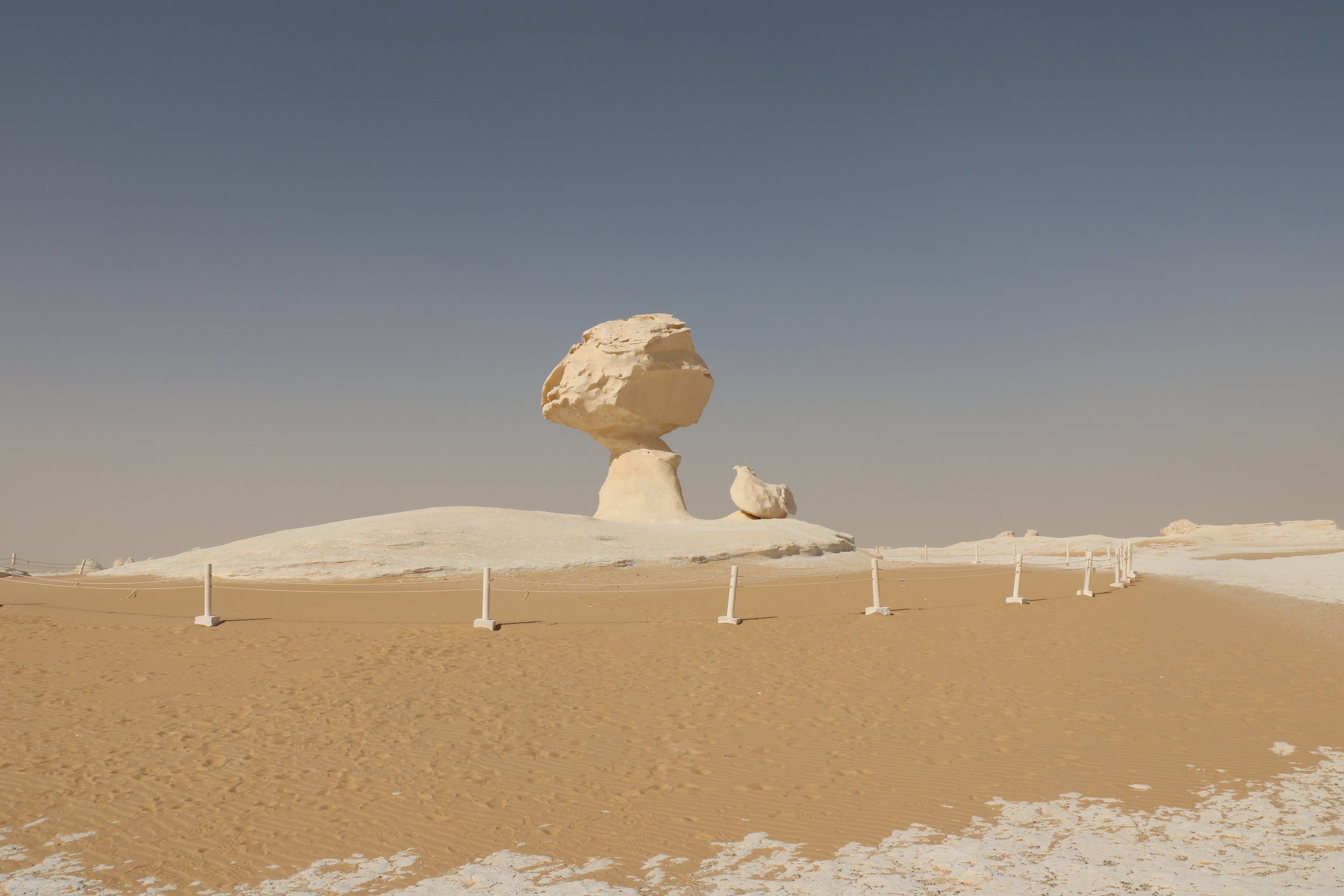 A desert landscape with a large, sculpted rock formation resembling a mushroom, surrounded by a white barrier and a clear blue sky.