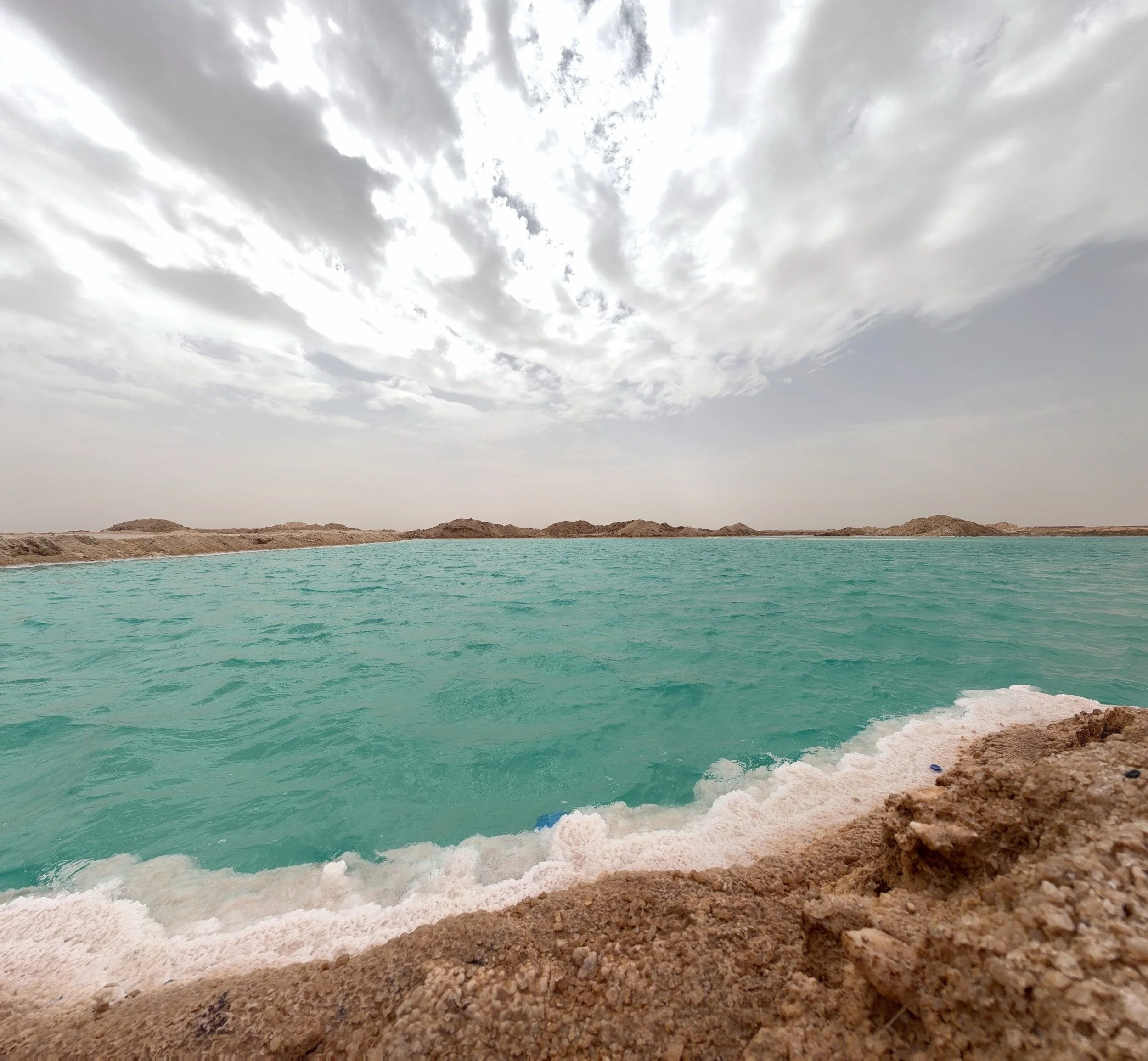 A desert landscape with turquoise water in the foreground and sandy hills in the background under a cloudy sky.