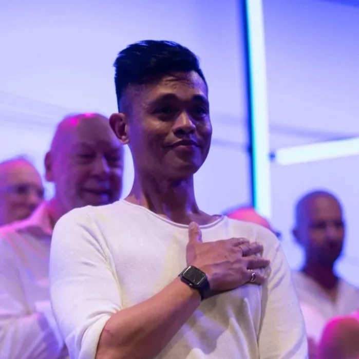 A young man with short dark hair making a pledge with his right hand over his chest in a room with purple and pink lighting. Other individuals are visible in the background.