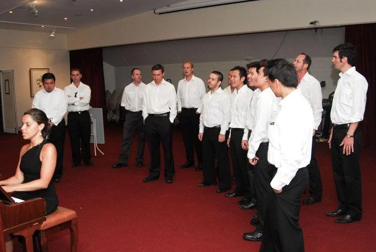 A woman playing a piano with a group of men in white shirts and black pants standing in line behind her, in a room with red carpet and dark red curtains.
