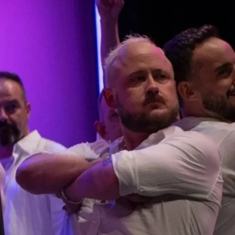 Three men standing close together in a dark room with purple lighting, with one in the center crossing his arms and appearing to be deep in thought.