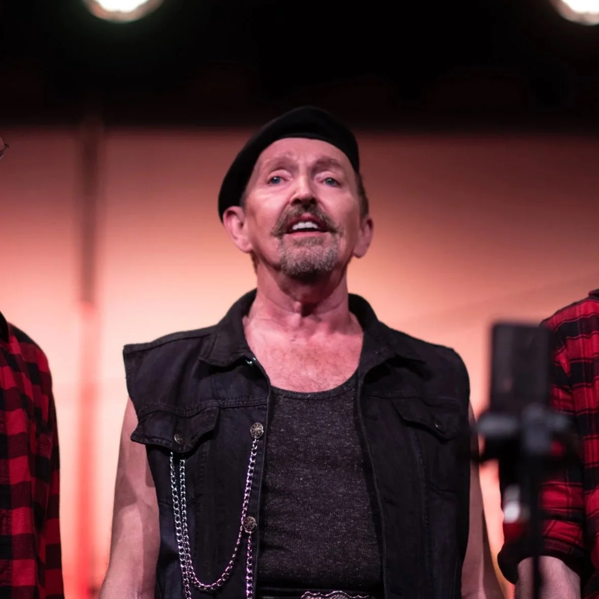 A man wearing a black sleeveless jacket, a dark shirt, a black beret, and a chain on his shoulder, standing confidently with a slight smile, in front of a blurred background with warm tones.