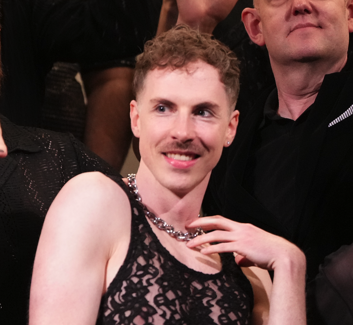 A person with short curly hair, wearing a black lace top and a silver chain necklace, smiling and looking to the side, surrounded by other people.