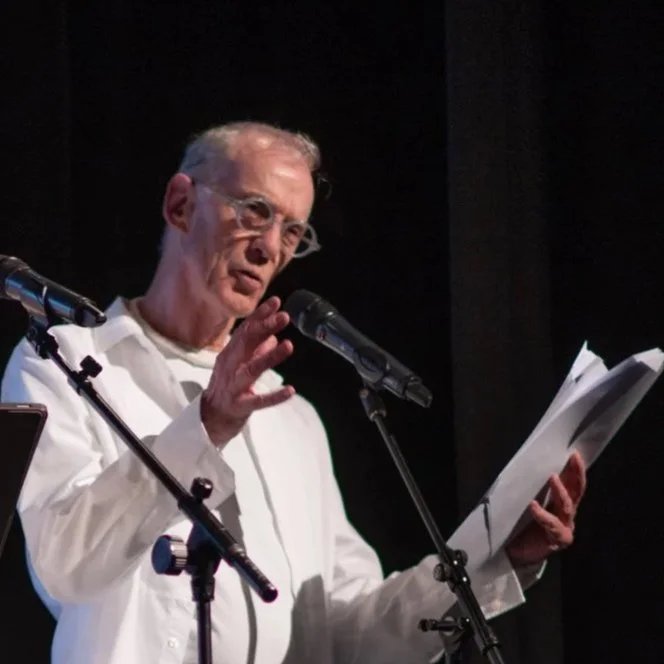 Older man with glasses, wearing a white shirt, speaking or reading into a microphone while holding papers on stage.